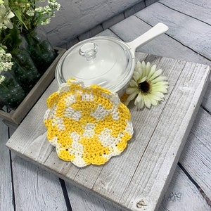 K&ouml;nnte beinhalten: Ein handgefertigter, gelb-wei&szlig;er Topflappen aus H&auml;kelarbeit mit gewelltem Rand. Eine transparente Glaspfanne mit wei&szlig;em Griff und eine wei&szlig;e Blume liegen auf einer verwitterten, grauen Holzoberfl&auml;che. Dekorative Blumen in gr&uuml;nen Glasflaschen im Hintergrund.