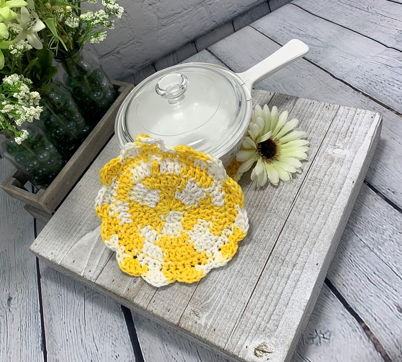 K&ouml;nnte beinhalten: Ein handgefertigter, gelb-wei&szlig;er Topflappen aus H&auml;kelarbeit mit gewelltem Rand. Eine transparente Glaspfanne mit wei&szlig;em Griff und eine wei&szlig;e Blume liegen auf einer verwitterten, grauen Holzoberfl&auml;che. Dekorative Blumen in gr&uuml;nen Glasflaschen im Hintergrund.