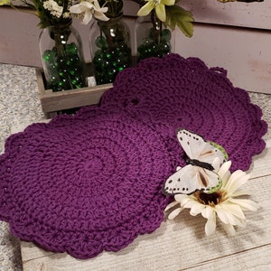 May include: Two purple crocheted round doilies with scalloped edges. The doilies are on a wooden surface with a white butterfly and a white flower.