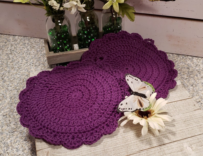 May include: Two purple crocheted round doilies with scalloped edges. The doilies are on a wooden surface with a white butterfly and a white flower.