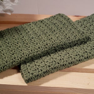 May include: Two green crocheted dishcloths or hand towels. The cloths are folded and stacked on top of each other on a wooden surface.