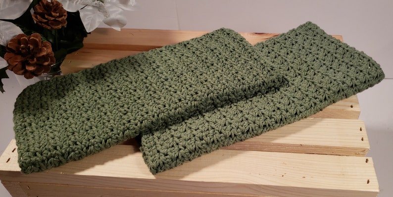 May include: Two green crocheted dishcloths or hand towels. The cloths are folded and stacked on top of each other on a wooden surface.