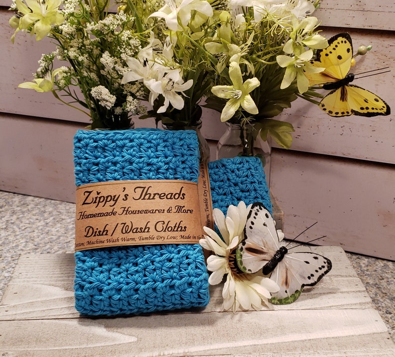 Puede incluir: Pa&ntilde;os de cocina de ganchillo turquesa con una etiqueta marr&oacute;n que dice "Zippy's Threads Homemade Housewares & More Dish/Wash Cloths". La imagen incluye flores blancas y mariposas.
