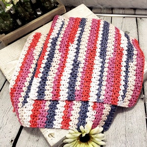 Pu&ograve; includere: Una coperta a righe all'uncinetto in tonalit&agrave; di rosa, rosso, bianco e grigio. La coperta &egrave; piegata a met&agrave; e si trova su una superficie di legno.