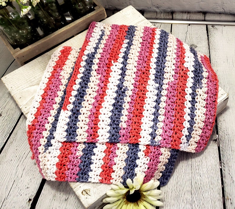 Pu&ograve; includere: Una coperta a righe all'uncinetto in tonalit&agrave; di rosa, rosso, bianco e grigio. La coperta &egrave; piegata a met&agrave; e si trova su una superficie di legno.
