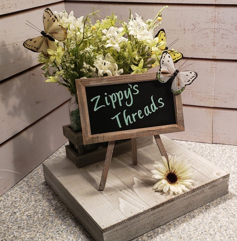 Puede incluir: Una exhibici&oacute;n r&uacute;stica de madera con una peque&ntilde;a pizarra que dice "Zippy's Threads". El letrero est&aacute; apoyado en un caballete en miniatura, con un ramo de flores blancas y decoraciones de mariposas. Una sola margarita blanca descansa en la exhibici&oacute;n.