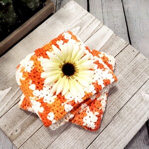 May include: Two square crochet coasters in orange and white, with a large cream-colored daisy on top. The coasters are handmade and have a textured appearance. The background is a rustic, weathered wooden surface.