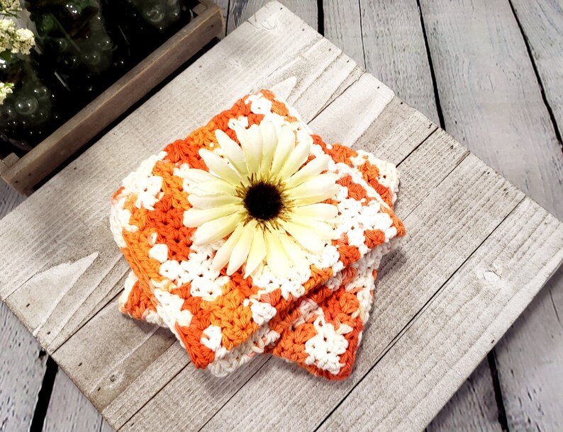 May include: Two square crochet coasters in orange and white, with a large cream-colored daisy on top. The coasters are handmade and have a textured appearance. The background is a rustic, weathered wooden surface.