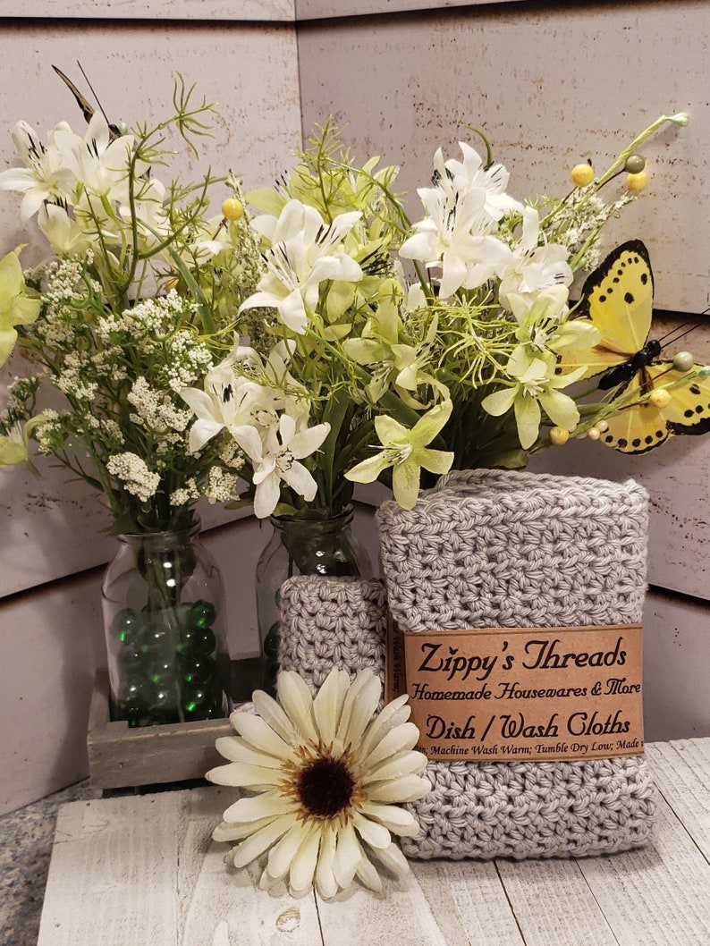 K&ouml;nnte beinhalten: Handgemachte, graue Sp&uuml;lt&uuml;cher mit dem Etikett "Zippy's Threads" werden zusammen mit einer wei&szlig;en G&auml;nsebl&uuml;mchen und einer Vase mit wei&szlig;en Blumen pr&auml;sentiert. Ein gelber Schmetterling schm&uuml;ckt die Anordnung.