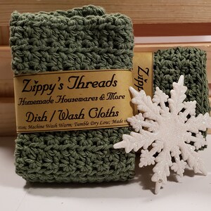 May include: Two green crocheted dishcloths with a white snowflake ornament. The dishcloths are labeled "Zippy's Threads Homemade Housewares & More Dish/Wash Cloths".