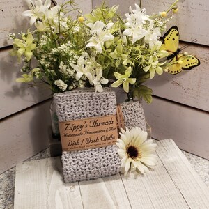 K&ouml;nnte beinhalten: Zwei geh&auml;kelte graue Geschirrt&uuml;cher mit einem braunen Papieretikett mit der Aufschrift "Zippy's Threads Dish/Wash Cloths". Die T&uuml;cher werden mit einem Strau&szlig; wei&szlig;er und gr&uuml;ner Blumen und einer gelben Schmetterlingsdekoration pr&auml;sentiert.