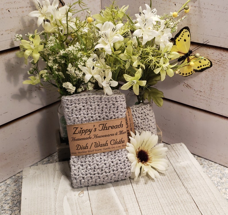 K&ouml;nnte beinhalten: Zwei geh&auml;kelte graue Geschirrt&uuml;cher mit einem braunen Papieretikett mit der Aufschrift "Zippy's Threads Dish/Wash Cloths". Die T&uuml;cher werden mit einem Strau&szlig; wei&szlig;er und gr&uuml;ner Blumen und einer gelben Schmetterlingsdekoration pr&auml;sentiert.