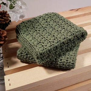 May include: A green crocheted dishcloth or washcloth. The cloth is folded and sitting on a wooden surface.