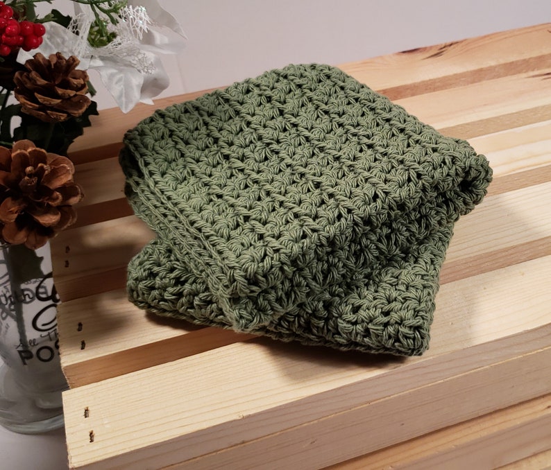 May include: A green crocheted dishcloth or washcloth. The cloth is folded and sitting on a wooden surface.