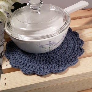 Pode incluir: Um pegador de panela azul de croch&ecirc; com borda ondulada suporta uma assadeira branca com tampa de vidro. O prato tem um design floral. O pegador de panela est&aacute; em uma superf&iacute;cie de madeira.