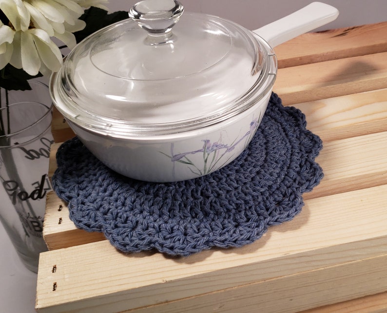 Pode incluir: Um pegador de panela azul de croch&ecirc; com borda ondulada suporta uma assadeira branca com tampa de vidro. O prato tem um design floral. O pegador de panela est&aacute; em uma superf&iacute;cie de madeira.