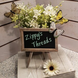 K&ouml;nnte beinhalten: Ein kleines Tafelschild auf einer Holzstaffelei zeigt den Text "Zippy's Threads". Das Schild ist von einem Blumenarrangement mit wei&szlig;en und gr&uuml;nen Blumen und dekorativen Schmetterlingen umgeben.