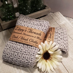 K&ouml;nnte beinhalten: Zwei graue geh&auml;kelte Geschirrt&uuml;cher mit einem braunen Papierband mit der Aufschrift "Zippy's Threads Homemade Housewares & More Dish/Wash Cloths". Eine wei&szlig;e Blume mit gelbem Zentrum liegt neben den T&uuml;chern. Die T&uuml;cher liegen auf einer hellen Holzoberfl&auml;che.