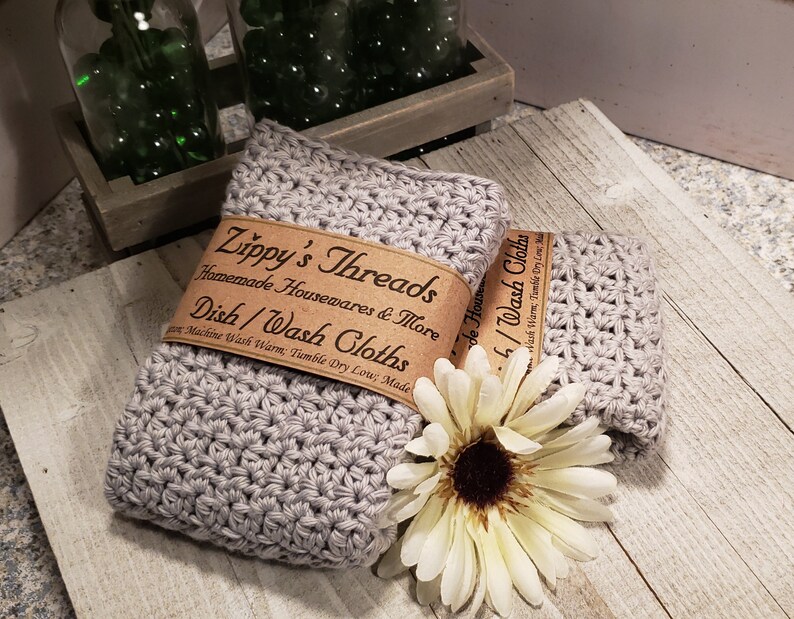 K&ouml;nnte beinhalten: Zwei graue geh&auml;kelte Geschirrt&uuml;cher mit einem braunen Papierband mit der Aufschrift "Zippy's Threads Homemade Housewares & More Dish/Wash Cloths". Eine wei&szlig;e Blume mit gelbem Zentrum liegt neben den T&uuml;chern. Die T&uuml;cher liegen auf einer hellen Holzoberfl&auml;che.