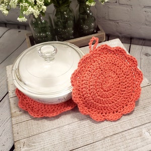 May include: Two coral-colored, crocheted pot holders with a scalloped edge. One pot holder is under a clear glass casserole dish with a lid. The other pot holder is next to the dish. The pot holders have a loop for hanging.