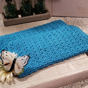 Puede incluir: Un objeto rectangular de color azul turquesa, tejido a mano, posiblemente un pa&ntilde;o o una peque&ntilde;a manta, descansa sobre una superficie de madera envejecida. Una mariposa blanca decorativa con manchas negras y una flor blanca est&aacute;n colocadas encima.