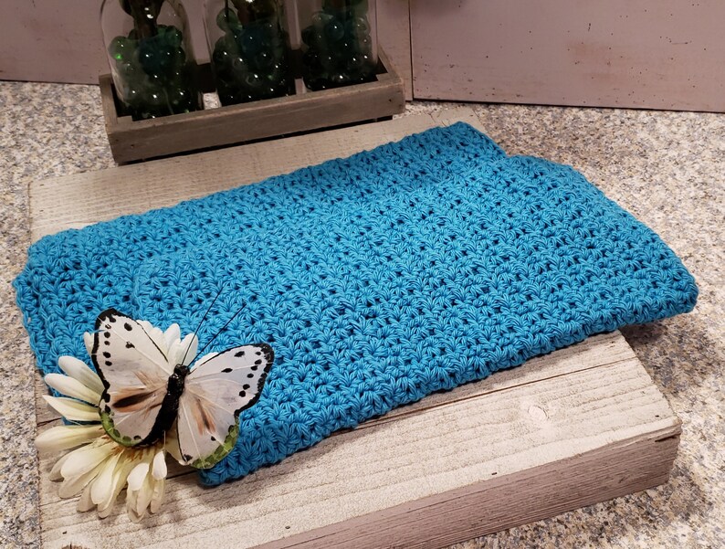 Puede incluir: Un objeto rectangular de color azul turquesa, tejido a mano, posiblemente un pa&ntilde;o o una peque&ntilde;a manta, descansa sobre una superficie de madera envejecida. Una mariposa blanca decorativa con manchas negras y una flor blanca est&aacute;n colocadas encima.
