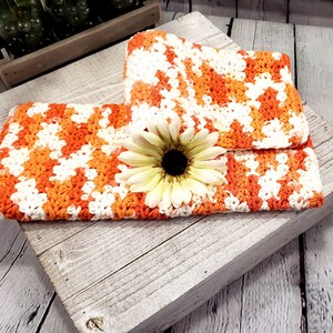 May include: Handmade crochet clutch in a vibrant orange, white, and cream color scheme. The clutch is folded over and features a large, cream-colored flower with a dark center. The crochet pattern is visible, and the clutch is resting on a rustic wooden surface.