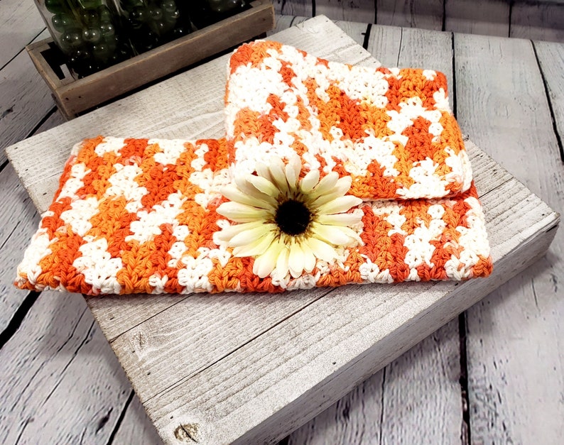 May include: Handmade crochet clutch in a vibrant orange, white, and cream color scheme. The clutch is folded over and features a large, cream-colored flower with a dark center. The crochet pattern is visible, and the clutch is resting on a rustic wooden surface.