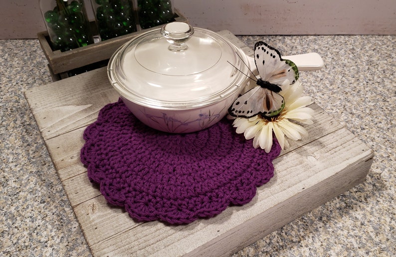 May include: Purple crocheted pot holder with a white ceramic pot with a glass lid on top. A white butterfly with black markings is perched on a white flower.