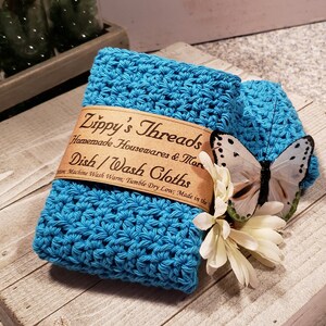 Puede incluir: Una pila de dos pa&ntilde;os de cocina de ganchillo turquesa con una mariposa y una flor decorativas. Los pa&ntilde;os de cocina tienen una etiqueta que dice "Zippy's Threads Homemade Housewares & More Dish/Wash Cloths".