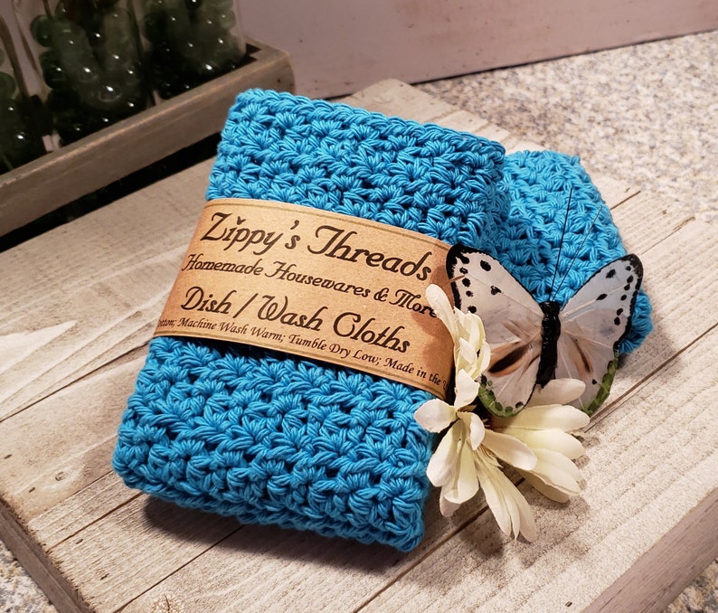 Puede incluir: Una pila de dos pa&ntilde;os de cocina de ganchillo turquesa con una mariposa y una flor decorativas. Los pa&ntilde;os de cocina tienen una etiqueta que dice "Zippy's Threads Homemade Housewares & More Dish/Wash Cloths".