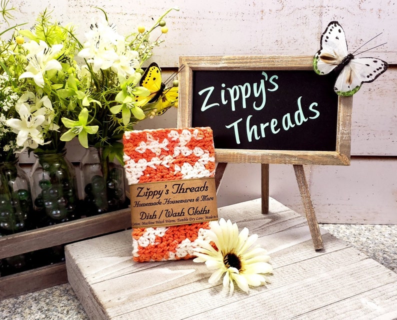 May include: An orange and white crocheted dishcloth is displayed with a small chalkboard sign that reads "Zippy's Threads". The image includes a wooden crate with flowers and a decorative butterfly.