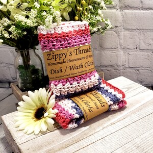 Pu&ograve; includere: Due strofinacci da cucina in crochet a righe rosa, viola e grigio con un'etichetta marrone che recita "Zippy's Threads Homemade Housewares & More Dish/Wash Cloths Cotton: Machine Wash Warm; Tumble Dry Low; Made in the USA"