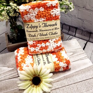May include: Two handmade crochet dish cloths in orange and white, stacked on top of each other. The cloths are labeled "Zippy's Threads" and "Dish/Wash Cloths". A white and yellow flower is in the foreground.