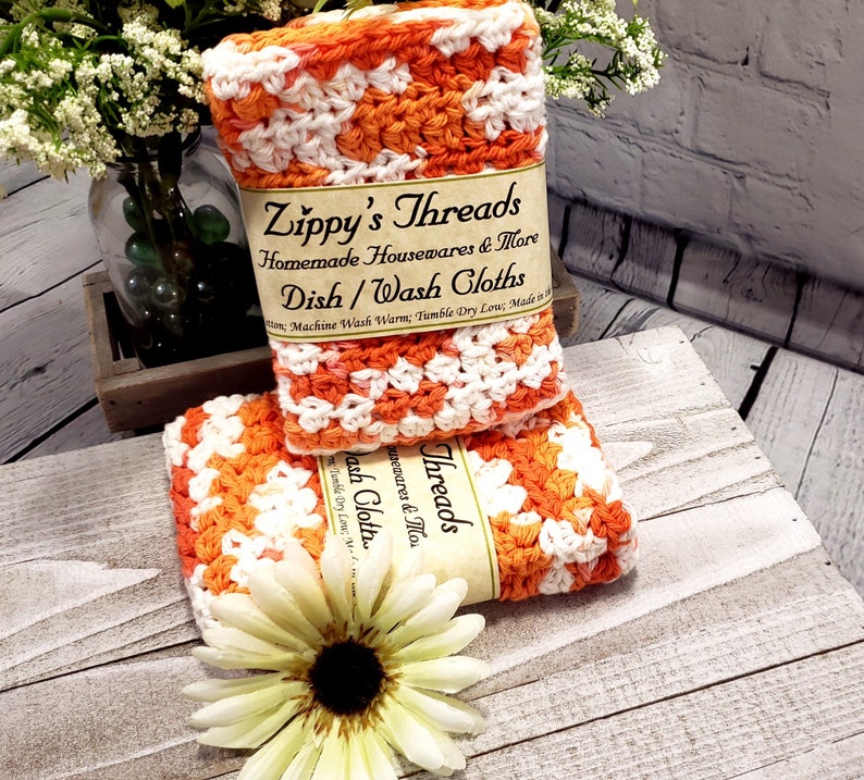 May include: Two handmade crochet dish cloths in orange and white, stacked on top of each other. The cloths are labeled "Zippy's Threads" and "Dish/Wash Cloths". A white and yellow flower is in the foreground.