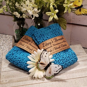 Puede incluir: Dos pa&ntilde;os de cocina de ganchillo turquesa con etiquetas marrones que dicen "Zippy's Threads Homemade Housewares Dish/Wash Cloths". Una margarita blanca y una mariposa est&aacute;n frente a los pa&ntilde;os. Flores y vegetaci&oacute;n en el fondo.