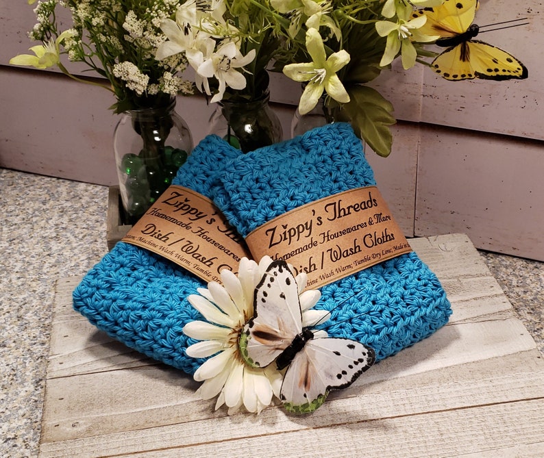 Puede incluir: Dos pa&ntilde;os de cocina de ganchillo turquesa con etiquetas marrones que dicen "Zippy's Threads Homemade Housewares Dish/Wash Cloths". Una margarita blanca y una mariposa est&aacute;n frente a los pa&ntilde;os. Flores y vegetaci&oacute;n en el fondo.