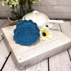 May include: A teal blue, crocheted hot pad with a scalloped edge. The circular pad is placed on a distressed white wooden surface, next to a white pot and a yellow and brown flower. The background includes white flowers in glass bottles.