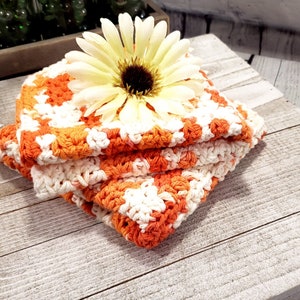 May include: A stack of three handmade crochet washcloths in orange and white, topped with a large yellow and brown flower. The washcloths are folded neatly on a rustic, weathered wooden surface.
