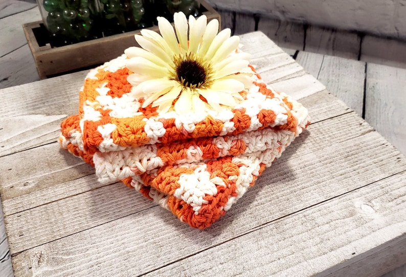 May include: A stack of three handmade crochet washcloths in orange and white, topped with a large yellow and brown flower. The washcloths are folded neatly on a rustic, weathered wooden surface.
