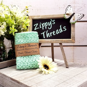 May include: A green crocheted dishcloth with a brown label that reads "Zippy's Threads Homemade Housewares & More Dish/Wash Cloths". The dishcloth is sitting on a white wooden surface with a small easel holding a black chalkboard sign that reads "Zippy's Threads".