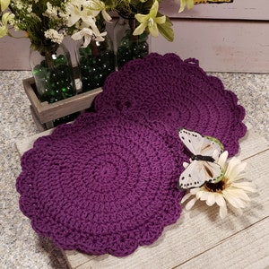 May include: Two purple crocheted pot holders with a scalloped edge. A white butterfly with black markings is perched on a white flower.