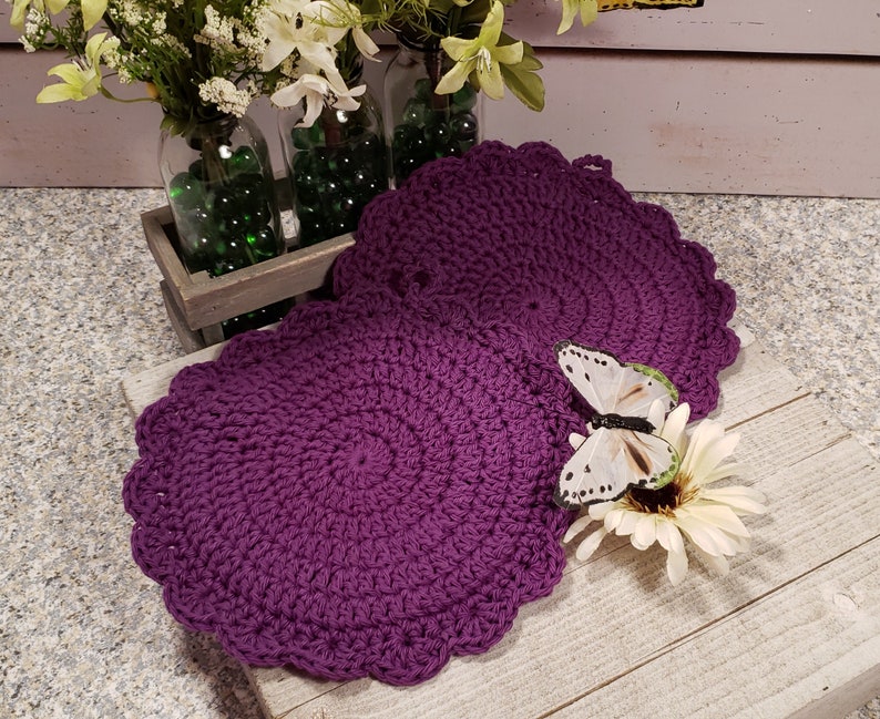 May include: Two purple crocheted pot holders with a scalloped edge. A white butterfly with black markings is perched on a white flower.