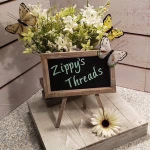 Pu&ograve; includere: Un piccolo cavalletto di legno con un cartello di ardesia che recita "Zippy's Threads". Il cavalletto &egrave; decorato con un arrangiamento di fiori bianchi e tre farfalle gialle.