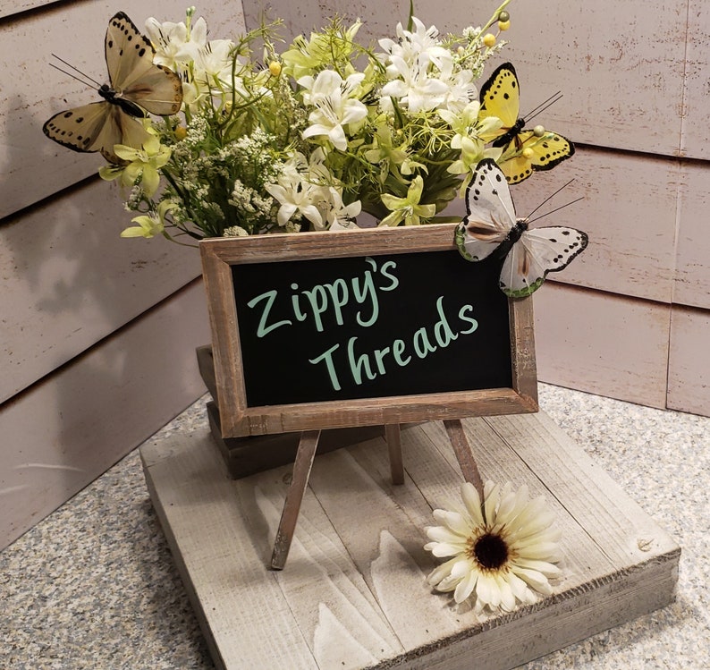 Pu&ograve; includere: Un piccolo cavalletto di legno con un cartello di ardesia che recita "Zippy's Threads". Il cavalletto &egrave; decorato con un arrangiamento di fiori bianchi e tre farfalle gialle.