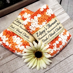 May include: Two handmade dish cloths in orange and white crochet, labeled "Zippy's Threads Dish/Wash Cloths." A white and yellow flower rests below the cloths on a rustic wooden surface. The cloths are made of a soft, absorbent material.