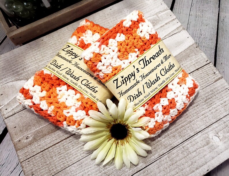 May include: Two handmade dish cloths in orange and white crochet, labeled "Zippy's Threads Dish/Wash Cloths." A white and yellow flower rests below the cloths on a rustic wooden surface. The cloths are made of a soft, absorbent material.