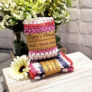 Pu&ograve; includere: Due strofinacci in crochet rosa, bianco e grigio con un'etichetta marrone che recita "Zippy's Threads Homemade Housewares & More Dish/Wash Cloths".