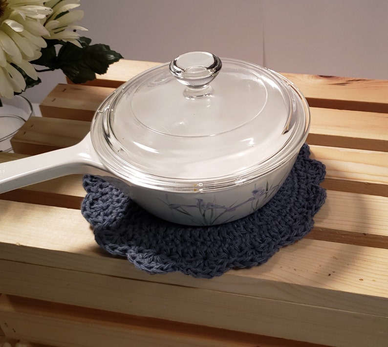 Pode incluir: Uma panela branca com tampa de vidro repousa sobre um descanso de panela azul de croch&ecirc; feito &agrave; m&atilde;o. O descanso de panela tem uma borda ondulada. A panela tem uma al&ccedil;a branca e um desenho floral na lateral.