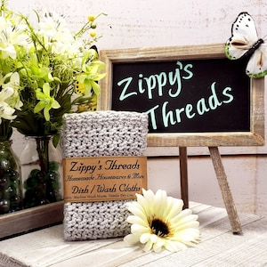 K&ouml;nnte beinhalten: Ein Stapel grauer geh&auml;kelter Geschirrt&uuml;cher mit einem braunen Etikett mit der Aufschrift "Zippy's Threads". Das Bild enth&auml;lt eine kleine Tafel mit den Worten "Zippy's Threads" und einen dekorativen Schmetterling. Ein Strau&szlig; wei&szlig;er und gr&uuml;ner Blumen befindet sich im Hintergrund.